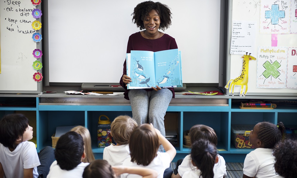 imagem de uma professora negra com um livro aberto nas mãos ensinando alunos em uma sala de aula